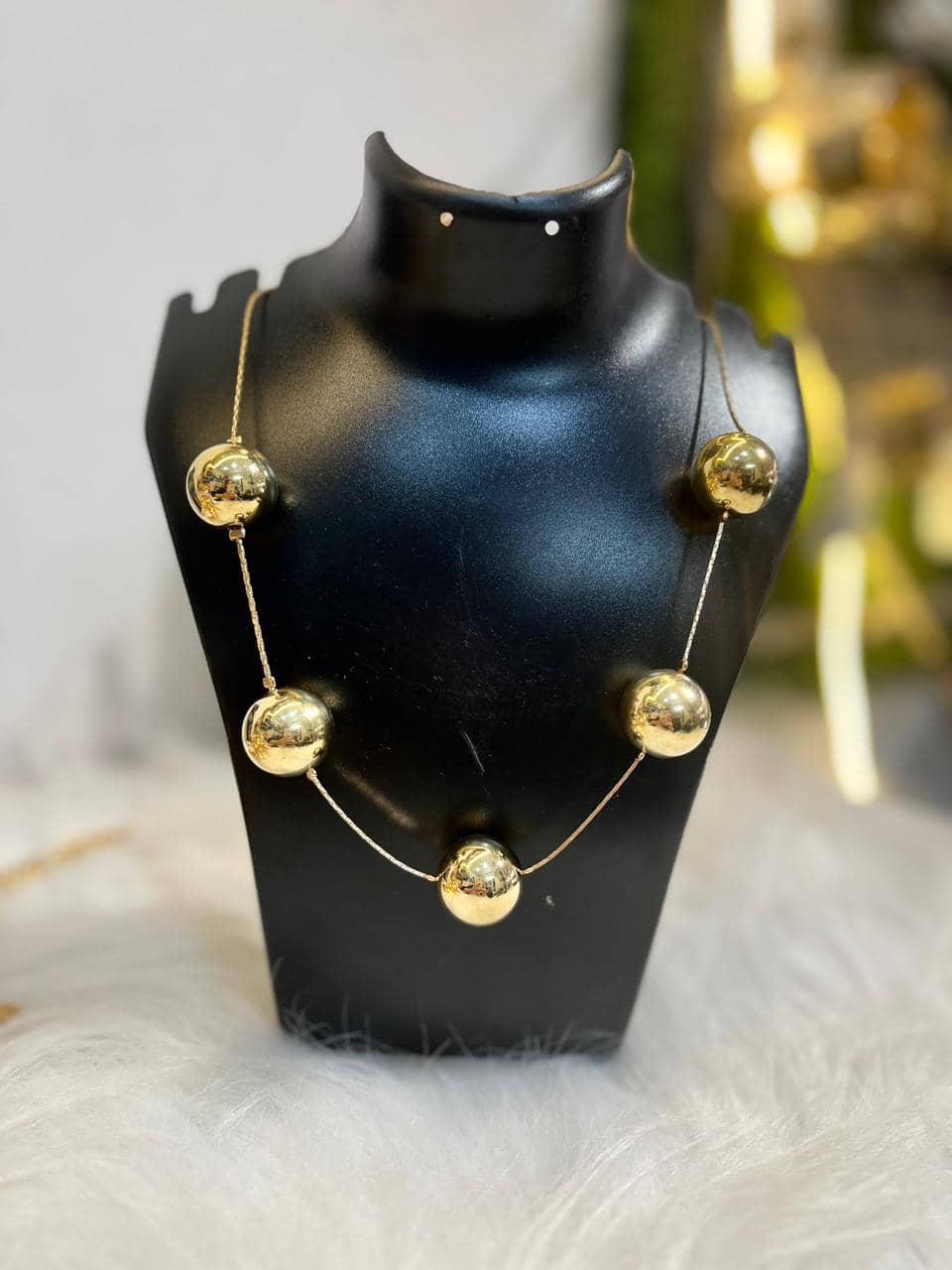 A modern necklace featuring five polished golden spheres on a delicate gold-tone wire, displayed on a black jewelry mannequin.